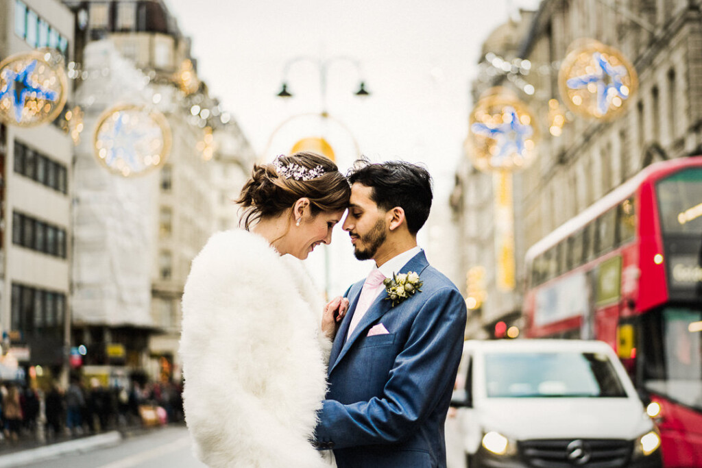 A smiling couple stands close together on a city street. The woman wears a white fur coat and the man is in a blue suit with a boutonniere. Decorative lights hang overhead, and a red double-decker bus is in the background.
