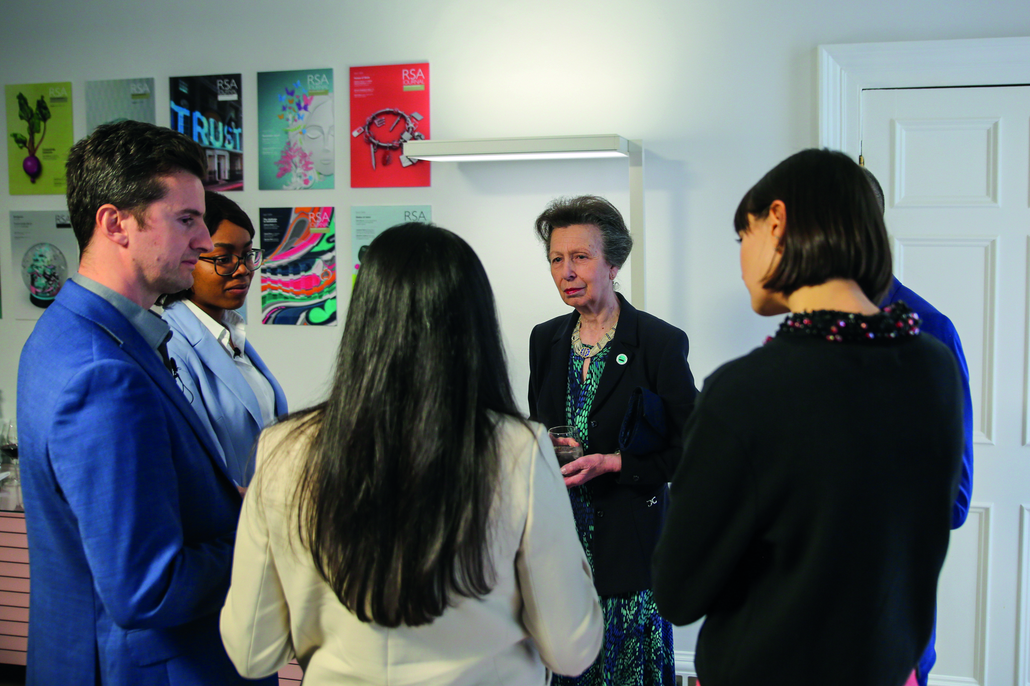 A group of five people engaged in conversation in a room with posters on the wall. One woman holding a glass appears to be speaking to the others, who are standing in a semi-circle. The atmosphere is business-casual.
