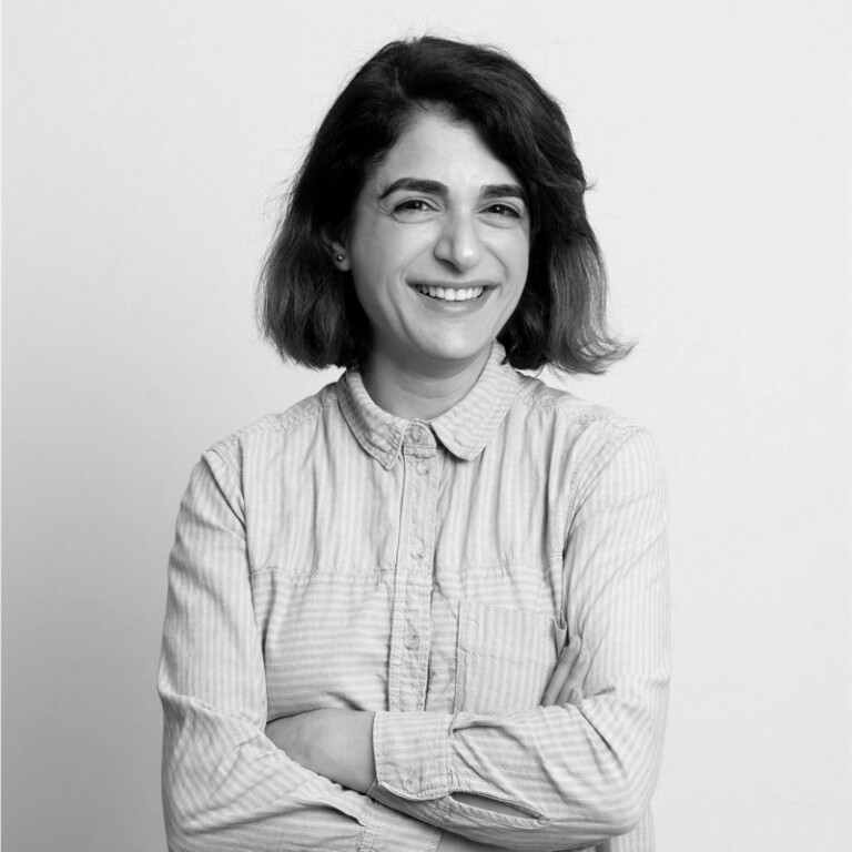 A person with shoulder-length hair smiles warmly at the camera. They are wearing a light-colored, button-up shirt with sleeves rolled up, and their arms are crossed. The background is plain white.
