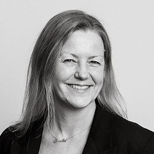 A black and white portrait of a person with long hair, smiling slightly, wearing a dark blazer and a necklace. The background is plain and light-colored.