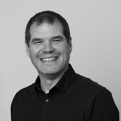 A black and white portrait of Jamie Cooke smiling warmly. They have short dark hair and are wearing a dark button-up shirt. The background is plain, highlighting their friendly expression.