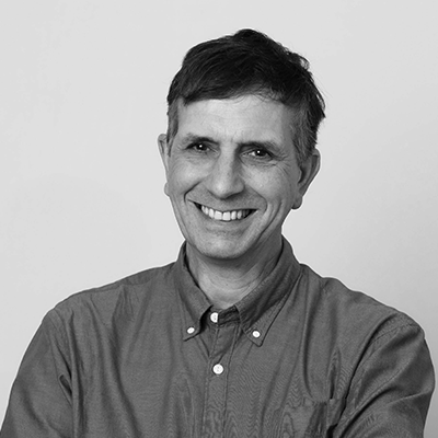 Smiling person in a button-up shirt with short, dark hair against a plain background. Image is in black and white.