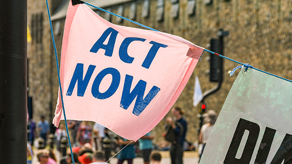 Protest scene with a pink banner reading "ACT NOW" in bold blue letters. The banner is attached to a blue string, with people and a building visible in the blurred background.