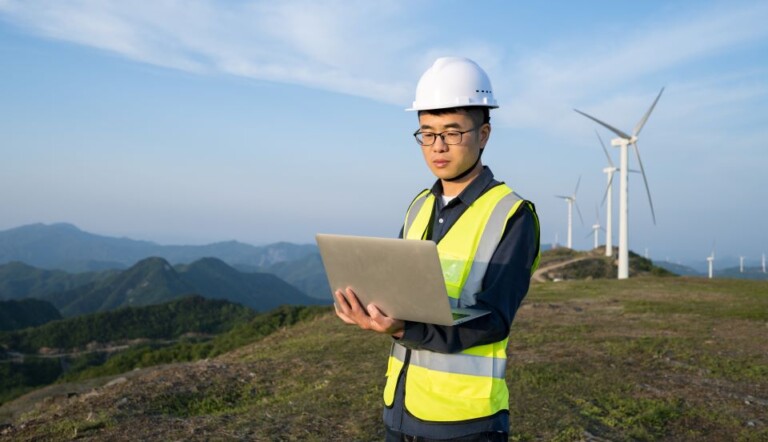 Person in a hard hat and safety vest using a laptop stands on a grassy hill with wind turbines and mountains in the background under a clear blue sky.