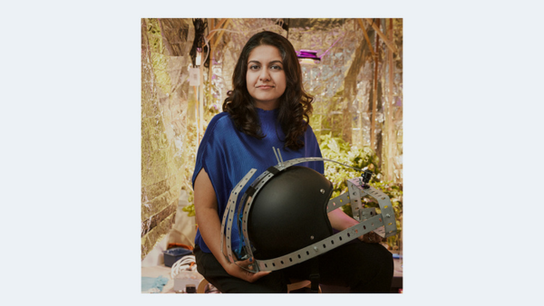 A woman sitting in a room with metallic foil insulation, holding a black helmet-like structure with metal brackets. She has long dark hair and is wearing a blue top. The background features plants and equipment.