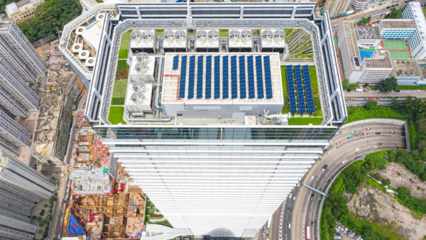 Aerial view of a tall building with a rooftop featuring solar panels and industrial equipment. Surrounding the building are busy roads, greenery, and other architectural structures in an urban landscape.