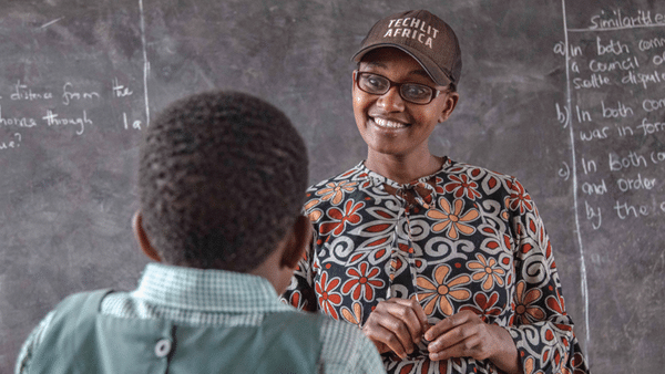 A teacher, wearing glasses and a floral patterned dress, smiles while talking to a student in a classroom. The teacher is wearing a cap with "Techlit Africa" written on it. There's writing on the blackboard behind them.