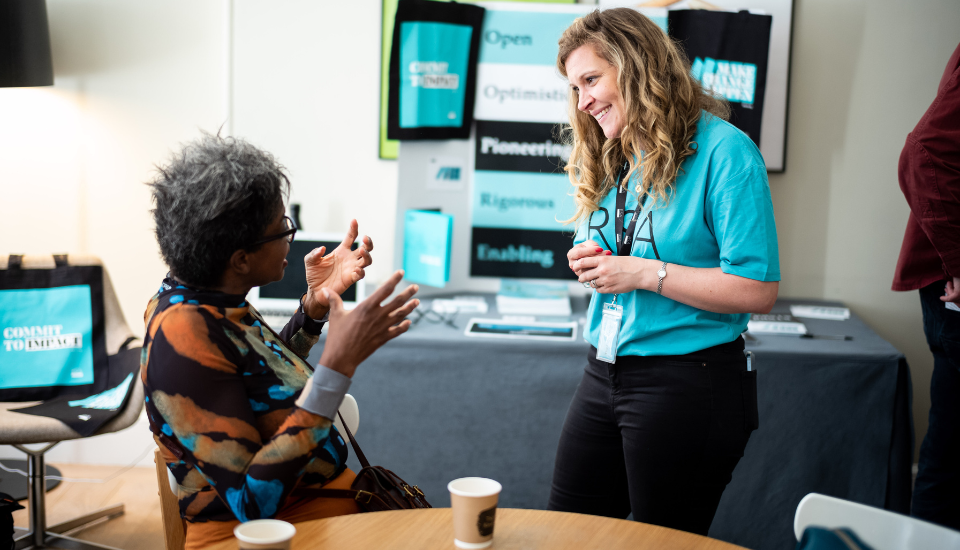 A person in a teal shirt and another seated individual engage in conversation at a table with coffee cups. A display with bags and promotional materials is visible in the background.