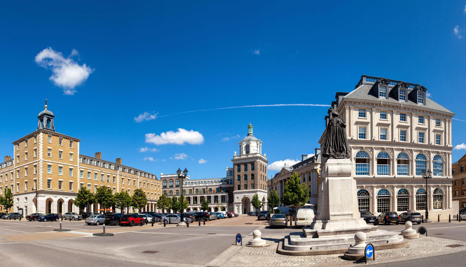 A spacious square with classical-style buildings under a clear blue sky. A statue is visible in the foreground, surrounded by cobblestone paving. Parked cars line the street, and trees dot the area, adding greenery.