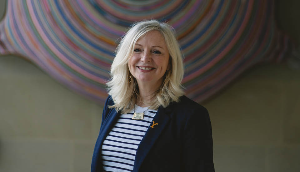 A person with long, blonde hair smiles at the camera. They are wearing a striped top and dark jacket, standing in front of a colorful, circular wall art piece.