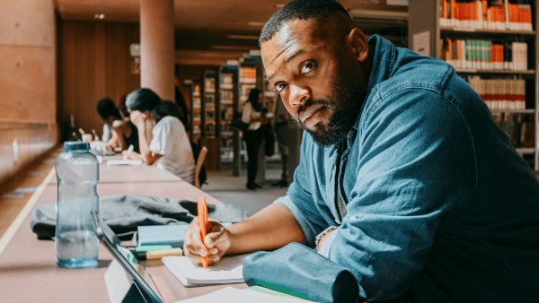 A man in a denim shirt sits at a library table, holding a pencil and surrounded by books and papers. He looks towards the camera, with shelves of books and other people studying in the background.