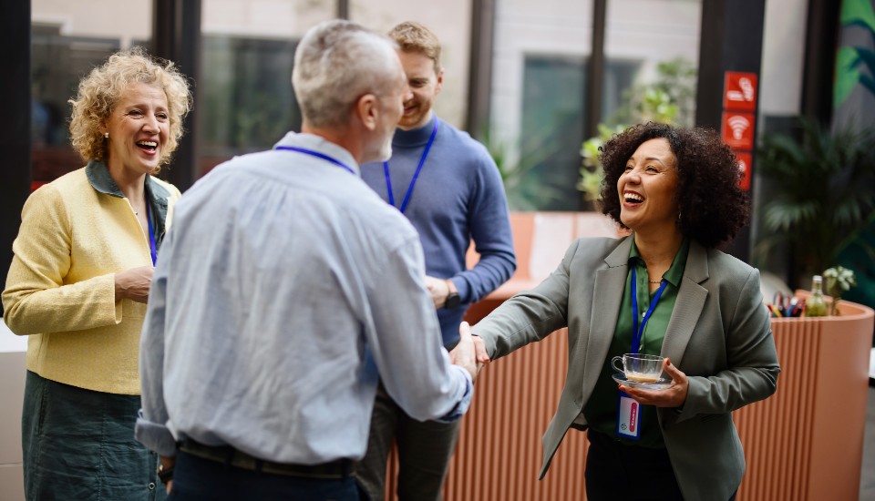Four people are gathered in a well-lit room. One person, a woman, is shaking hands with a man who has his back to the camera. Two others are smiling, observing the exchange. They all wear name tags and appear to be in a casual business setting.