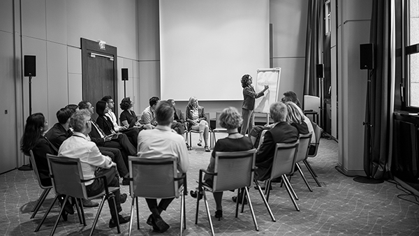A group of people sit in a circle of chairs in a conference room. One person stands and writes on a flip chart at the front. The setting is professional, with attendees focused on the speaker. The room has a large screen and several windows.