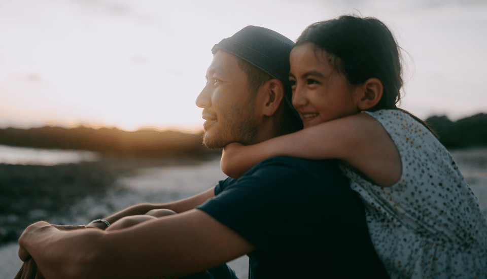 A man wearing a cap gives a piggyback ride to a smiling child. They are looking towards the horizon, enjoying a beach setting with a soft, warm sunset in the background.