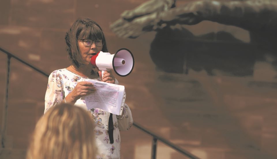 A person with glasses and a floral shirt speaks into a megaphone while holding notes. They stand outdoors against a blurred background with a staircase and a sculpture.