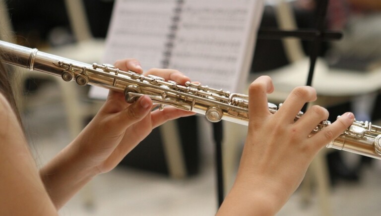 Close-up of a person's hands playing a silver flute, with fingers positioned on the keys. In the background, a music stand displays sheet music.