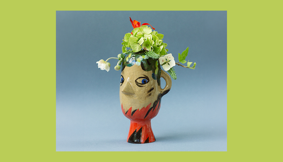 Colorful ceramic vase with a face design, featuring blue eyes and red accents. It holds a bouquet of green hydrangeas, white flowers, and greenery. The background is a soft blue with a bright green border.