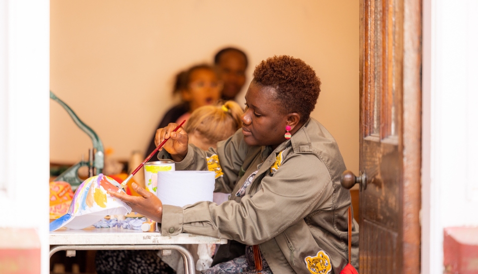 A person in a green jacket sits at a table, painting a white pumpkin with orange designs. A child with blonde hair is nearby, and blurred people are in the background, inside a room with an open door.
