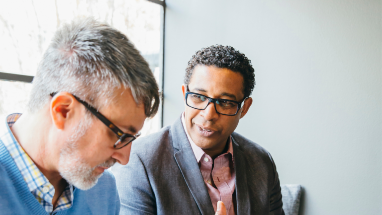 Two men sitting and talking. The man on the left has gray hair and glasses, wearing a blue sweater over a plaid shirt. The man on the right has curly hair and glasses, wearing a suit jacket over a red shirt. They're indoors with natural light coming in.