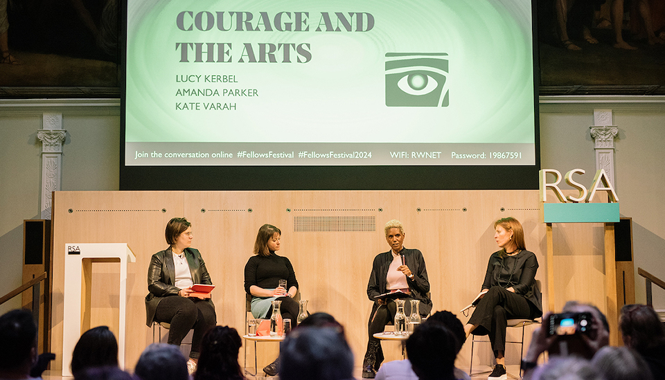 Four panelists sit on stage in discussion, with a large screen behind them displaying the event title "Courage and the Arts" and participant names. The audience listens attentively. The setting appears formal and professional.