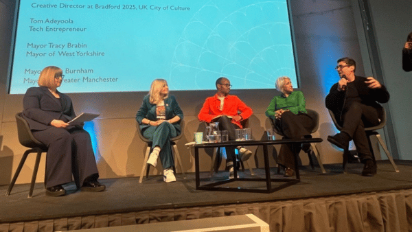Five people are seated on a stage panel. A woman in a red sweater speaks while others listen. Behind them is a blue screen with text showing names and titles. A row of empty glasses and cups is placed on a small table in front of them.
