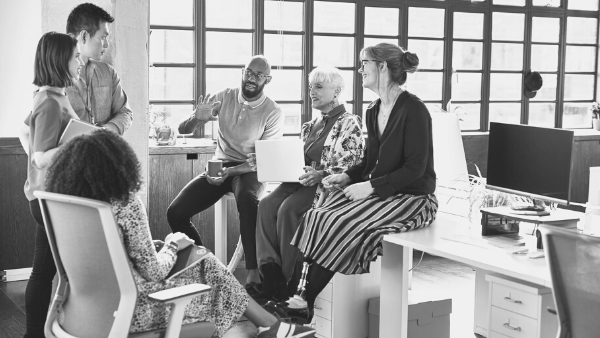 A diverse group of people in a casual office setting having a discussion. They are seated and standing around a desk, with large windows in the background, allowing natural light to fill the room.
