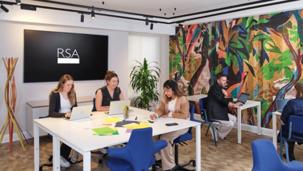 A modern office space with four people working on laptops at a table. Bright blue chairs and colorful paper are on the table. The wall features vibrant, abstract artwork and a large screen displaying "RSA.