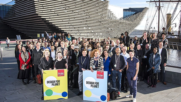 A large group of people stands outside a modern building with a ship in the background. They hold posters that say "Design for Planet." The crowd is diverse, and everyone is dressed casually. The atmosphere is bright and sunny.