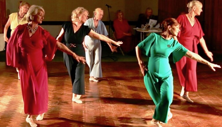 A group of older women in colorful dresses dancing gracefully on a wooden floor. They form a line, extending one arm forward and one leg back. In the background, a few people are seated near audio equipment.