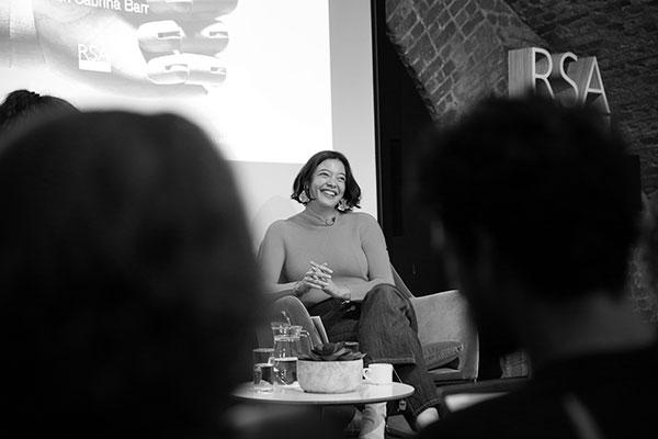 A woman sitting on a stage, smiling and speaking to an audience. She is wearing a long-sleeve top and pants. A screen and "RSA" logo are visible in the background. The image is in black and white. Audience members are partially visible in the foreground.