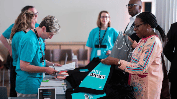 A woman in a colorful outfit receives a gift bag at a registration table, while others in teal shirts assist her. A group of people stands nearby, and folders labeled "MIT IMPACT" are visible on the table.