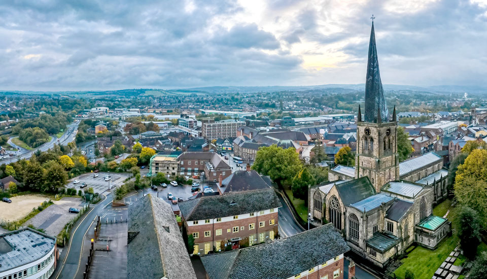 Aerial view of a large stone church with a tall spire in a town. Surrounding buildings and roads are visible, set against a backdrop of lush greenery and hills under a cloudy sky.
