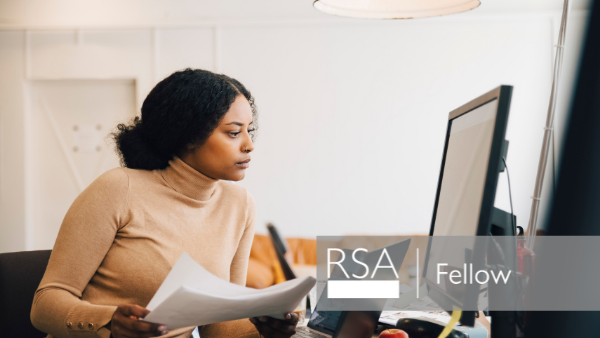 Woman in a beige turtleneck working at a desk, holding papers and looking intently at a computer screen. The words "RSA Fellow" are overlaid on the image. The setting appears to be a modern office.