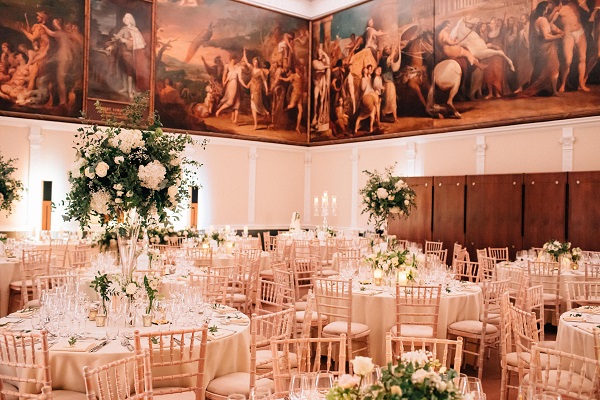 The elegant ballroom, perfect for an event at the RSA, features round tables with white and gold decor, high floral centerpieces, and wooden chairs. Large murals adorn the walls, depicting classical scenes while warm lighting enhances the luxurious ambiance.