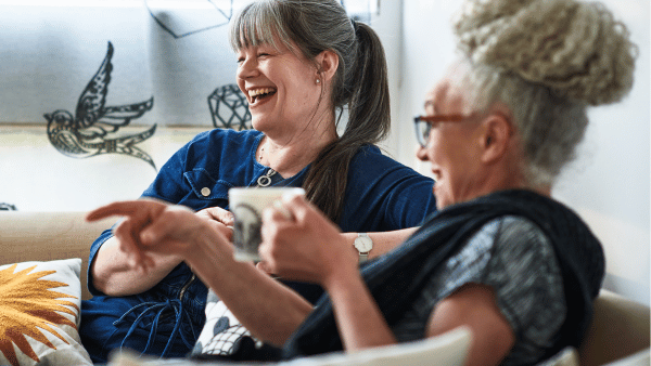 Two women sit on a couch, smiling and laughing. One points outward while holding a mug; the other holds a cup and wears glasses. Behind them is a window with decorative bird designs.