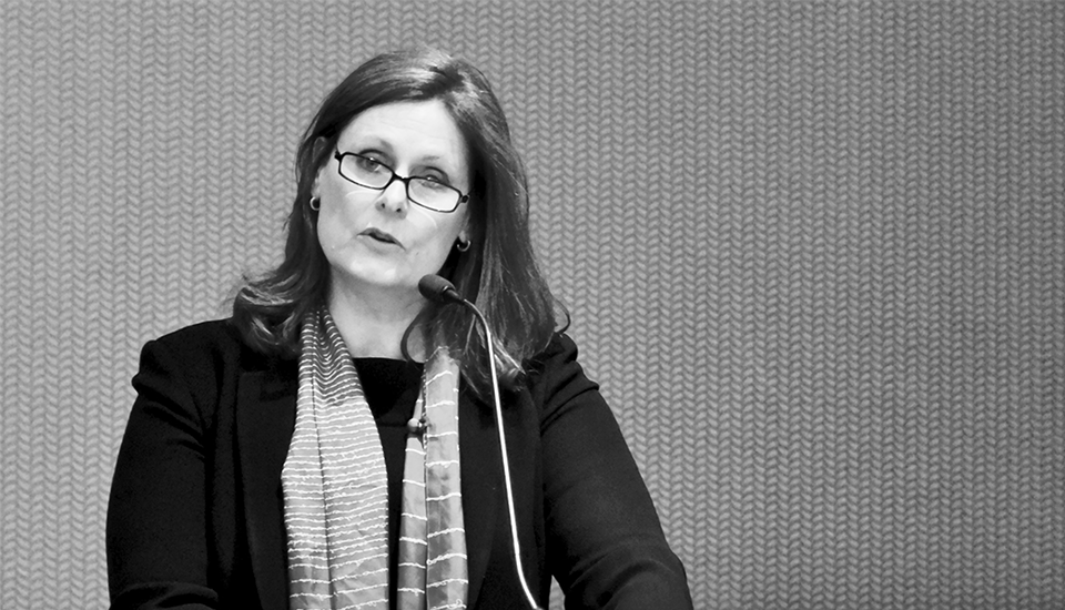 A woman with glasses speaks into a microphone. She wears a dark blazer and a striped scarf. The background is a textured wall. The image is in black and white.
