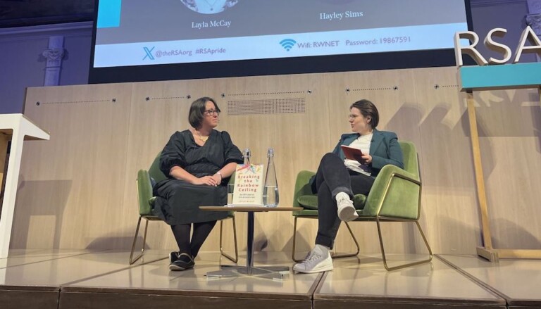 Two individuals sit on stage in armchairs engaged in discussion. Between them is a book titled "Breaking The Bamboo Ceiling." A screen behind them displays the names Layla McCay and Hayley Sims, along with event hashtags and a Wi-Fi password.