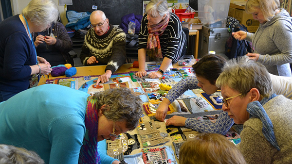 A group of older adults collaborates around a table, engaging in a crafting activity. They are focused on a large, colorful textile piece, surrounded by yarn and craft materials in a cozy indoor setting.