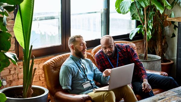 Two people sit on a leather couch in a modern, plant-filled office, focused on a laptop screen. One wears a blue shirt, the other a red plaid shirt. Large windows let in natural light.