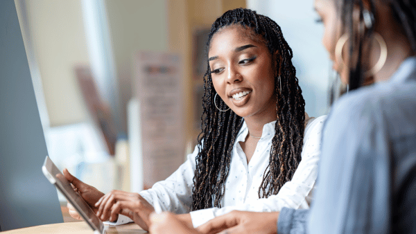 Two women are sitting at a table, engaged in conversation. One woman with braided hair is holding a tablet and smiling, while the other, blurred in the foreground, is listening. The setting appears to be an informal indoor space.