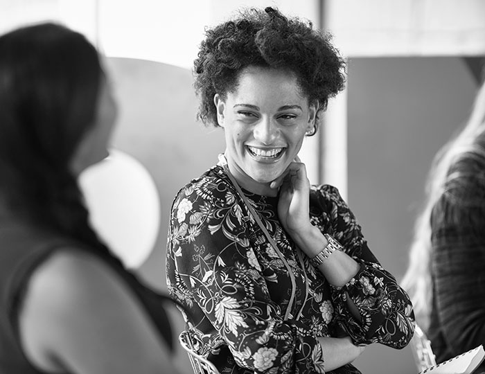A person with curly hair smiles warmly and looks to the left at another blurred person in the foreground. They are wearing a floral-patterned outfit. The setting appears to be indoors, with soft lighting. Black and white photo.