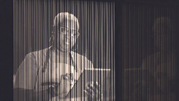 A monochromatic image of a person viewed through vertical strings or threads, holding a tablet or book. The lines give a blurred effect, slightly distorting the image. The person appears focused and is wearing glasses.