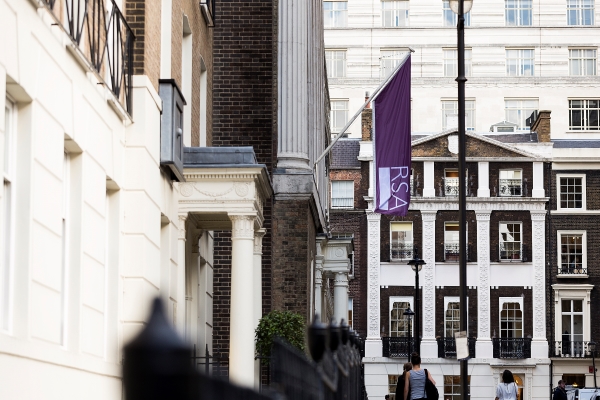 Street view showing a row of classic brick and stone buildings with a prominent purple flag labeled "RSA." A man and woman walk along the sidewalk, partially visible on the right.