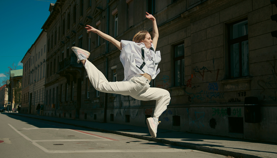 A person in white clothing and sneakers performs a dynamic leap on an empty urban street lined with old buildings. They are mid-air with arms and legs extended, showcasing grace and athleticism under a clear blue sky.