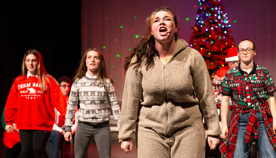 A person in a fuzzy costume passionately performs on stage with several others. They stand in front of a Christmas tree, and the group behind wears festive attire, including sweaters and Santa hats. Colorful stage lights illuminate the background.