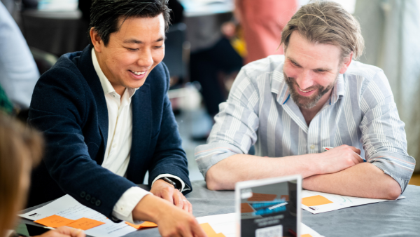 Two men sitting at a table, engaged in discussion. One is pointing at something on a paper with orange notes. Both are smiling, suggesting a positive interaction. The background is blurred with people and soft lighting.