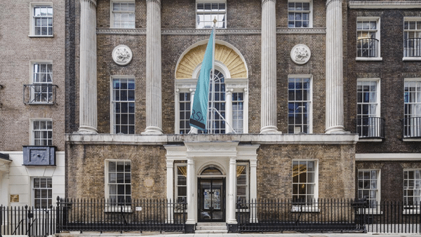 A historic building with brick facade, tall arched windows, and white columns flanking the entrance. A green flag is displayed above the door. A black wrought iron fence borders the front.