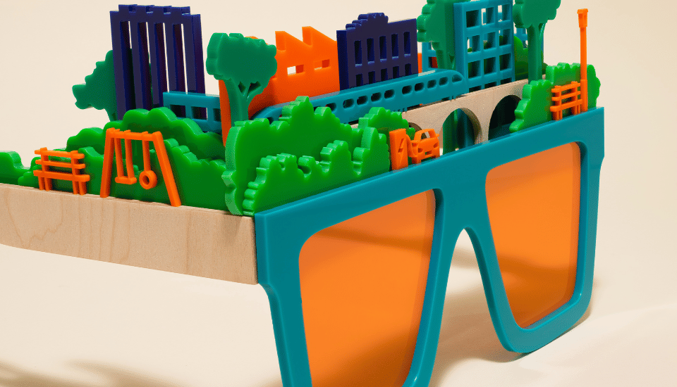 Quirky oversized glasses with orange lenses adorned with a playful miniature model of a cityscape, featuring colorful buildings, trees, a bridge, and small park elements on top.