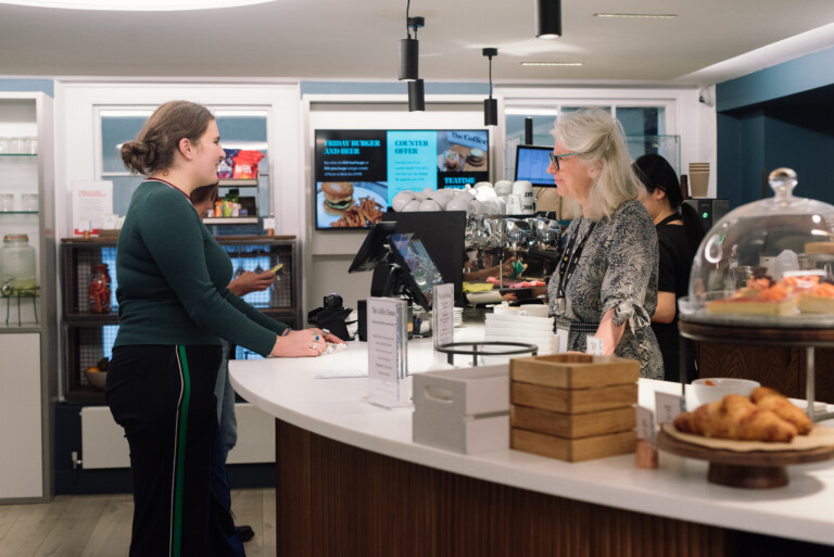 A woman with a ponytail and a green sweater is standing at a café counter speaking to a barista with grey hair and glasses. The café is bright with various baked goods displayed and digital menus in the background.