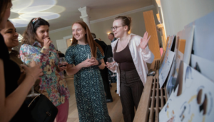 Four people stand in a room, engaged in conversation and looking at a display of artwork on a ledge. They are holding wine glasses, and the atmosphere seems casual and lively. The space is softly lit, adding warmth to the scene.